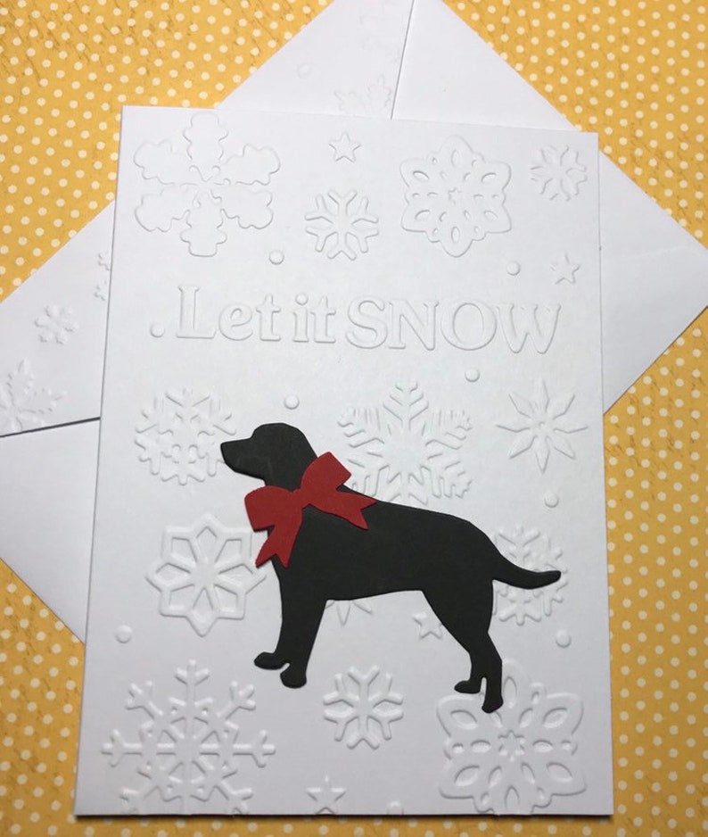 May include: White greeting card with embossed snowflakes and the phrase "Let it SNOW." A black dog with a red bow is featured on the card. Includes a white envelope. The background is yellow with white polka dots.
