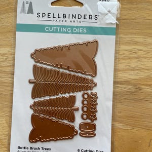 Puede incluir: Un paquete de troqueles de corte Spellbinders Paper Arts. El paquete contiene seis troqueles de corte de metal color cobre con forma de árboles de cepillo y elementos decorativos. El paquete está etiquetado como "Bottle Brush Trees".