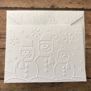May include: White embossed greeting card set featuring three snowmen wearing hats and scarves, with snowflakes in the background. The matching envelope also has embossed snowflakes. A winter-themed stationery set.