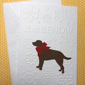 May include: White greeting card with embossed snowflakes and the phrase "Let it SNOW." A brown dog silhouette with a red bow is featured. Includes a matching envelope.