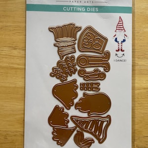 Puede incluir: Un conjunto de 13 formas troqueladas de metal dorado para manualidades. Las formas incluyen un gnomo, una piña, un sombrero y una bota. El paquete dice "Dancin' Gnome" y "Gnome Dansant".