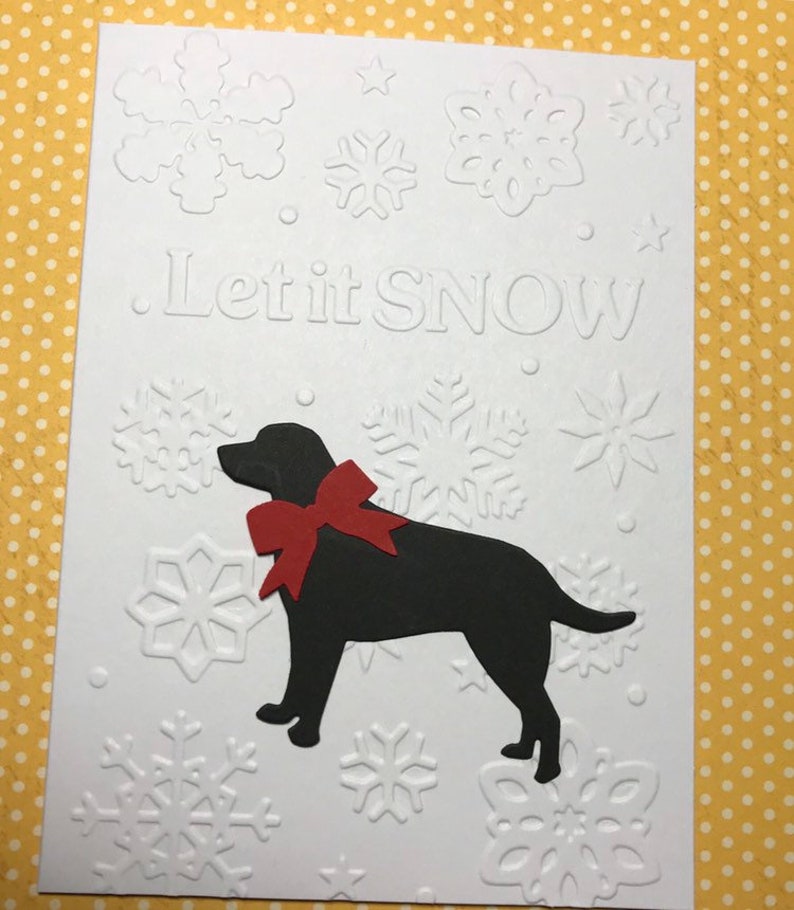 May include: White greeting card with embossed snowflakes and the phrase "Let it SNOW." A black silhouette of a dog with a red bow is featured on the card. The background is a yellow polka dot pattern.