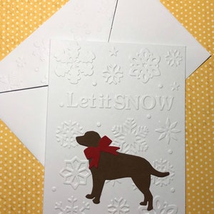 May include: A white embossed Christmas card with the words "Let it SNOW" and snowflake designs. A brown dog with a red bow is featured on the card. An accompanying white envelope is included.