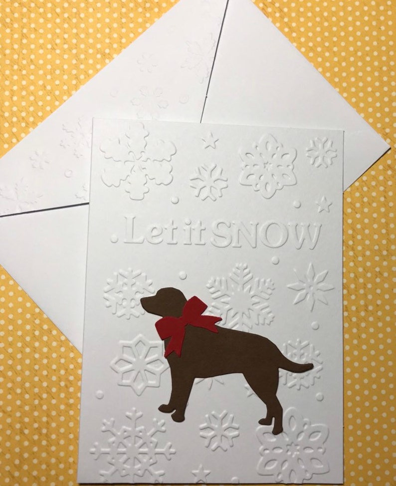 May include: A white embossed Christmas card with the words "Let it SNOW" and snowflake designs. A brown dog with a red bow is featured on the card. An accompanying white envelope is included.