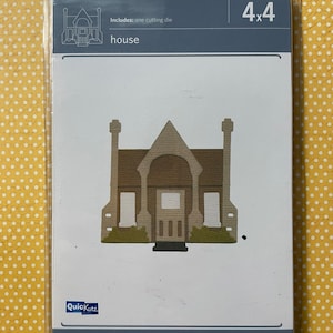 May include: A die-cutting template for a house, measuring 4x4 inches. The template is made of brown cardstock and features a detailed design of a house with a pointed roof, windows, and a door. The template is packaged in a clear plastic sleeve with the text "Quic Kutz" and "Includes: one cutting die" printed on it.