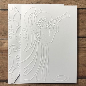 May include: White embossed greeting card featuring an angel playing a trumpet, with a halo and flowing gown. The card is paired with a matching envelope with a decorative embossed pattern. The design is simple and elegant.