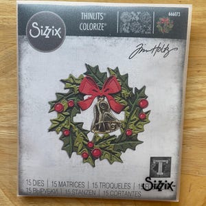 May include: A Sizzix Thinlits die set featuring a Christmas wreath design with a red bow and bell. The packaging includes the text "THINLITS COLORIZE" and "Jim Holtz". The set contains 15 dies.