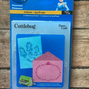 May include: A blue Disney Cuttlebug embossing folder set featuring Cinderella. The package includes two embossing folders with princess designs and the text "emboss gaufrage". The set also includes a pink envelope and a blue card.