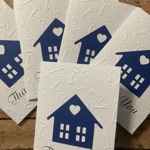 May include: A stack of white thank you cards with a blue house design and a heart on the front. The cards have a white embossed pattern of keys on the background. The text "Thank You" is written in black cursive on the front of the card.