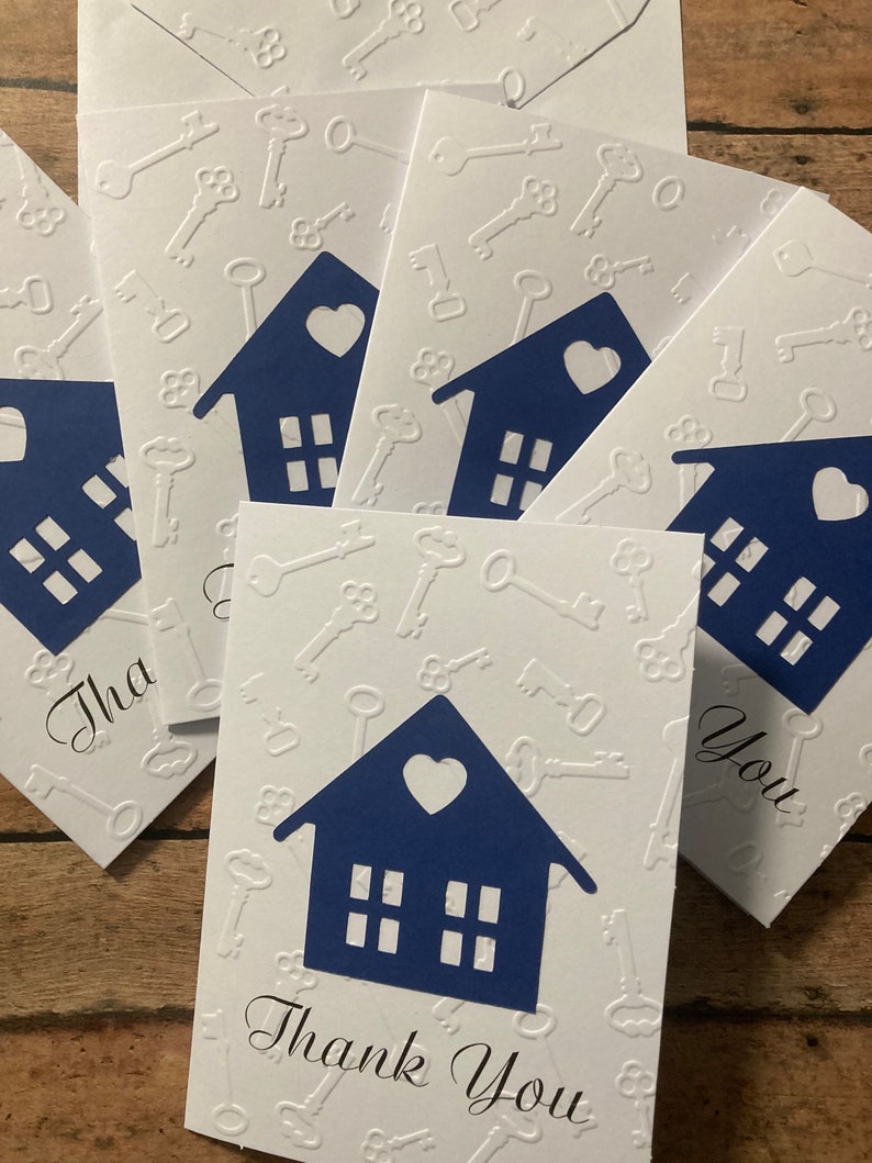 May include: A stack of white thank you cards with a blue house design and a heart on the front. The cards have a white embossed pattern of keys on the background. The text "Thank You" is written in black cursive on the front of the card.