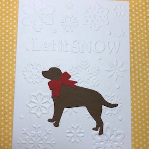 May include: A white greeting card with embossed snowflakes and the phrase "Let it SNOW". A brown dog silhouette with a red bow is featured. The card has a festive, winter theme.