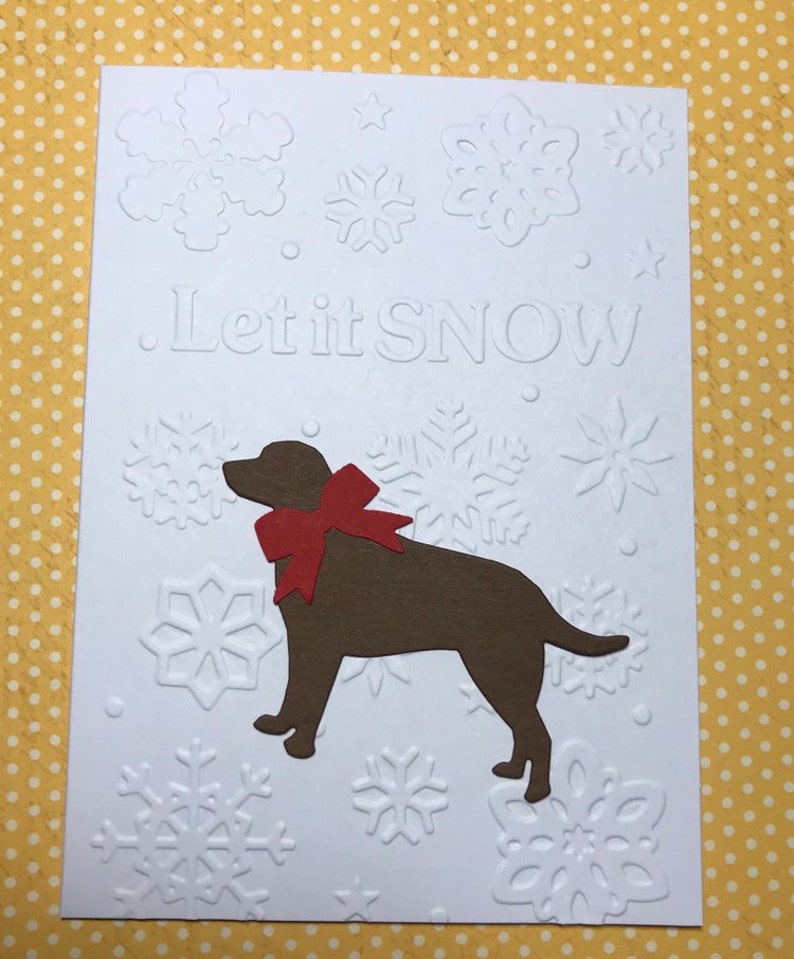 May include: A white greeting card with embossed snowflakes and the phrase "Let it SNOW". A brown dog silhouette with a red bow is featured. The card has a festive, winter theme.
