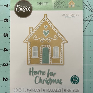 May include: A die-cut metal template for a gingerbread house, with the words "Home for Christmas" in a teal blue color. The template is designed to be used with a die-cutting machine. The template is part of a set of 6 dies.