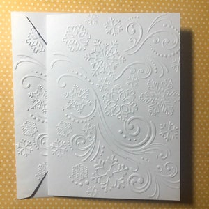 May include: A white greeting card and envelope set with an embossed snowflake and swirl design. The card features a detailed pattern of snowflakes and flowing lines, creating a textured, wintery aesthetic. The envelope also has a matching embossed snowflake design.