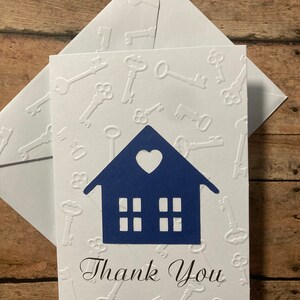 May include: A white card with a blue house silhouette and a heart cutout. The house has four windows and the card is embossed with a pattern of keys. The card says "Thank You" in black cursive writing.