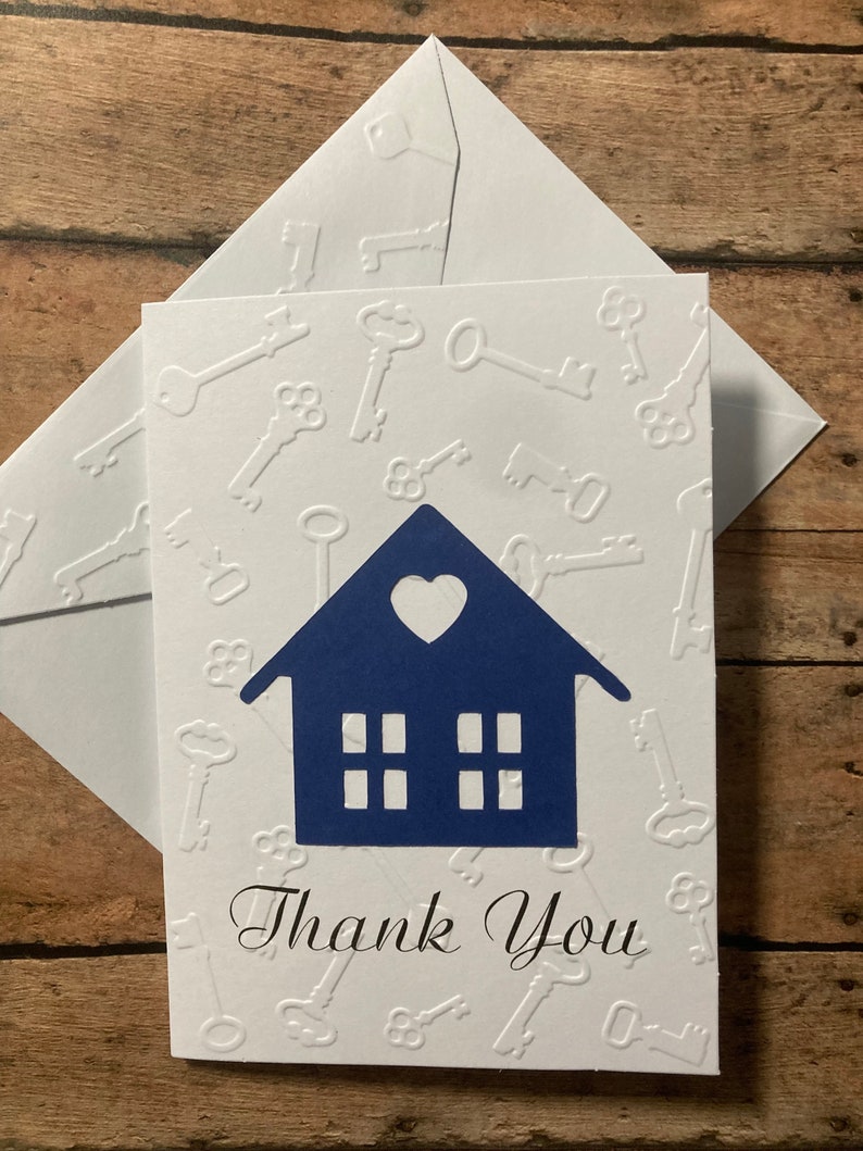 May include: A white card with a blue house silhouette and a heart cutout. The house has four windows and the card is embossed with a pattern of keys. The card says "Thank You" in black cursive writing.