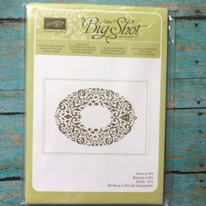 May include: A Stampin' Up! Sizzix Big Shot Textured Impressions embossing folder in a light green package. The product features a holiday frame design with an oval wreath pattern of intricate swirls, stars, and small trees in the center.