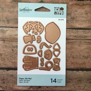 May include: A set of 14 metal die-cut templates for crafting. The templates are in the shape of a tiger, a hat, a bow tie, and other accessories. The templates are gold in color and are packaged on a white card with the Spellbinders logo and the product name "Tiger, Oh My!"