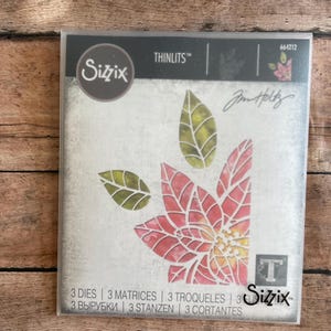 May include: A Sizzix Thinlits die set featuring a poinsettia flower and leaves design. The package includes the text "3 DIES | 3 MATRICES | 3 TROQUELES" and the Sizzix logo. The design is red, yellow, and green.