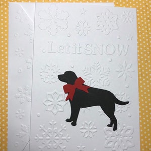 May include: White greeting card with embossed snowflakes and the words "Let it SNOW." A black silhouette of a dog with a red bow is featured. The card is paired with a white envelope, perfect for winter greetings.