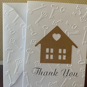 May include: A white card with a brown paper house cutout with a heart-shaped window and three windows below. The card is decorated with a white embossed pattern of keys. The text "Thank You" is written in black cursive lettering.