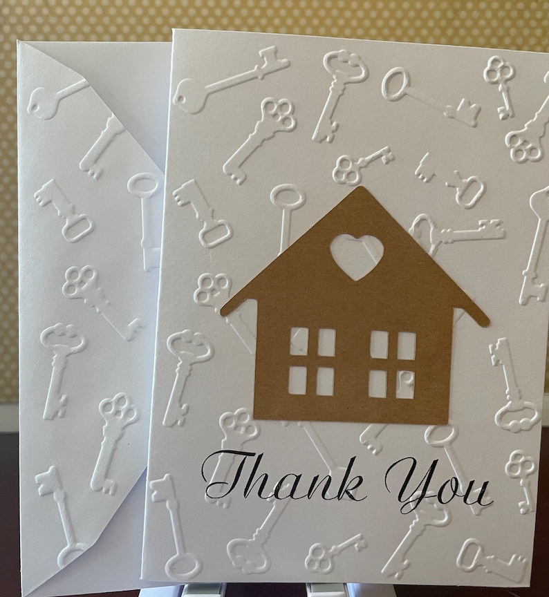 May include: A white card with a brown paper house cutout with a heart-shaped window and three windows below. The card is decorated with a white embossed pattern of keys. The text "Thank You" is written in black cursive lettering.