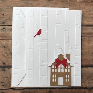 May include: White greeting card with embossed birch tree design, a red cardinal, and a brown gingerbread house with a red bow. The card is on a wooden surface.