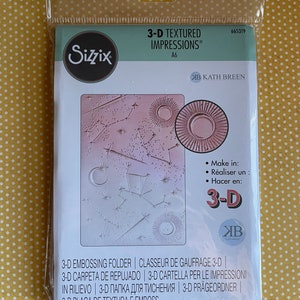 May include: A white and pink 3-D embossing folder with a celestial design. The folder is labeled "3-D Textured Impressions" and "Kath Breen".