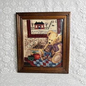 May include: A framed cross-stitch embroidery featuring a large golden teddy bear holding a smaller blue teddy bear. The scene includes a red house, a basket of berries, a red apple, and the alphabet letters A through S on a cream background.
