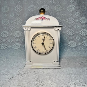 May include: A white ceramic mantel clock with a gold-tone finial on top. The clock face features black Roman numerals and ornate hands. A floral decal with pink and purple flowers is centered above the clock face. The frame has column details.