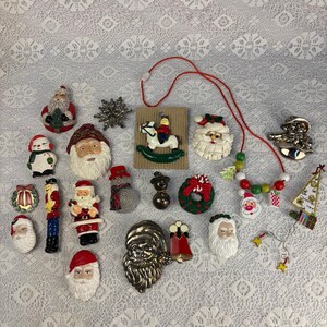 Puede incluir: Una colección de broches y adornos navideños que incluye varias caras de Papá Noel, un muñeco de nieve, un cascanueces, un caballito balancín en una tarjeta, una corona, una campana, una estrella metálica y un collar de cuentas con colgantes.