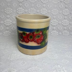 May include: A cylindrical ceramic crock with a cream-coloured base and two horizontal navy blue stripes. The centre features a hand-painted design of bright red strawberries with green leaves and white seeds, encircling the exterior of the vessel.