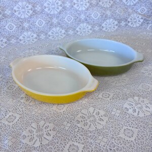 May include: Two oval-shaped baking dishes with integrated handles. One dish is bright yellow on the exterior with a white interior, while the other dish is olive green on the exterior with a white interior. Both dishes have smooth, glossy surfaces.