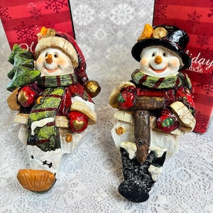 May include: Two snowman figurines wearing red and green winter clothing. The left snowman holds a green pine tree, while the right snowman holds a brown shovel. Both figures feature orange carrot noses, black hats or caps, and red gloves.