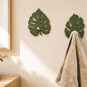 Könnte beinhalten: Zwei dunkelgrüne Wandhaken in Form von Monstera-Blättern. Jeder Haken verfügt über einen zentralen hellen Holzstift zum Aufhängen von Gegenständen. Ein Haken ist leer, während am anderen ein gefaltetes beiges Handtuch hängt.