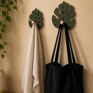 Könnte beinhalten: Zwei Wandhaken in Form von dunkel olivgrünen Monstera-Blättern mit hellen Holzstiften. Ein beiges Handtuch mit Fransen hängt am kleineren Haken, während eine schwarze Stofftasche am größeren Haken befestigt ist und an der Wand hängt.