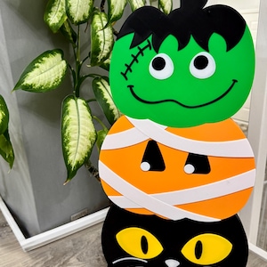 May include: A vertical stack of three decorative Halloween figures. The top is a green pumpkin with black hair and stitches. The middle is an orange pumpkin wrapped in white bandages. The bottom is a black cat with large yellow eyes and white whiskers.