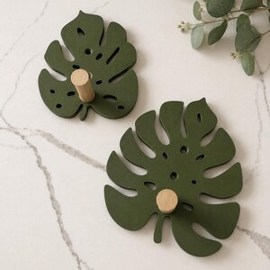 May include: Two wall hooks shaped like dark olive green monstera leaves. Each leaf-shaped base features a central light-colored wooden cylindrical peg extending outward. The design includes cut-out details within the leaf structure.