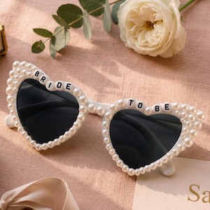 Puede incluir: Gafas de sol blancas en forma de corazón con lentes negras, adornadas con perlas de imitación. Las gafas de sol tienen las palabras "BRIDE" y "TO BE" en letras blancas. Un accesorio para una boda o evento nupcial.