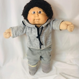 May include: A soft-sculpture doll with dark brown yarn hair, round cheeks, and a single visible tooth. The doll wears a heather gray hooded zip-up jacket with black drawstrings and matching gray pants with light yellow trim at the ankles.