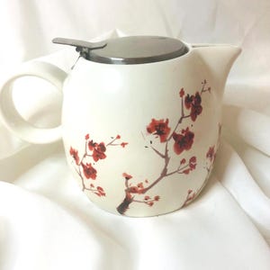 May include: A cream-colored ceramic teapot featuring a painted red and brown cherry blossom branch design. The teapot has a rounded body, a curved handle, a wide spout, and a circular metallic lid with a small handle tab on the top.