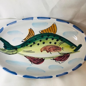May include: An oval ceramic platter featuring a hand-painted green and yellow fish with black spots, orange dorsal fins, and dark red lower fins. The rim is decorated with a repeating pattern of blue brushstrokes against a white background.