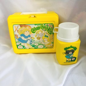 May include: A yellow plastic lunchbox and matching thermos set featuring Cabbage Patch Kids graphics. The lunchbox displays children gardening, while the thermos shows a child with a hoe. Both items feature the Cabbage Patch Kids logo.