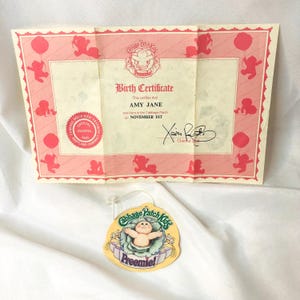May include: A Cabbage Patch Kids Preemie birth certificate for Amy Jane born November 1st, featuring a pink border with baby silhouettes and an official hospital seal, accompanied by a round yellow Cabbage Patch Kids Preemie hang tag.