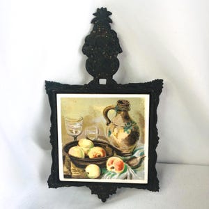 May include: A black cast iron trivet featuring an ornate handle and a square ceramic tile insert. The tile displays a still life painting of a ceramic pitcher, two clear glasses, and a bowl filled with yellow and peach-colored fruits.