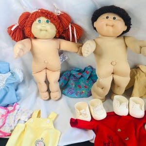 May include: Two Cabbage Patch Kids dolls with soft bodies and vinyl heads. One doll has orange yarn hair in pigtails with white ribbons and green eyes. The other doll has short brown yarn hair. Included are various doll clothes and two pairs of white shoes.