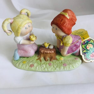 May include: A Cabbage Patch Kids porcelain figurine featuring two children kneeling on a green grassy base with small flowers. One child has blonde pigtails and holds a yellow chick, while the other has red hair and looks at a nest on a tree stump.