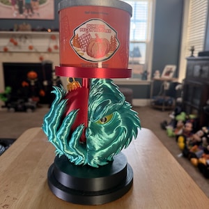May include: A candle warmer lamp featuring a base with a sculpted, teal-coloured Grinch character face and a red Christmas ornament. The lamp holds a cylindrical Bath & Body Works Pumpkin Pecan Waffles candle with an orange label and silver lid.