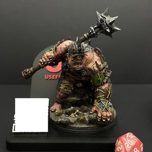 May include: A detailed miniature figure of a cyclops-like creature, holding a spiked mace. The figure is painted with various colors, including shades of brown, pink, and green. The figure is on a rocky base, with a red 20-sided die nearby.