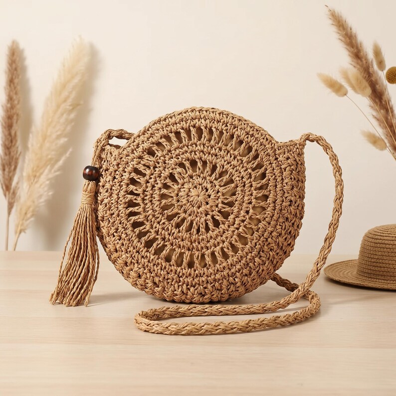May include: A round, tan woven straw crossbody bag featuring an intricate openwork circular pattern on the front. The bag includes a long braided shoulder strap and a decorative tassel accent attached to the side with a dark brown wooden bead.